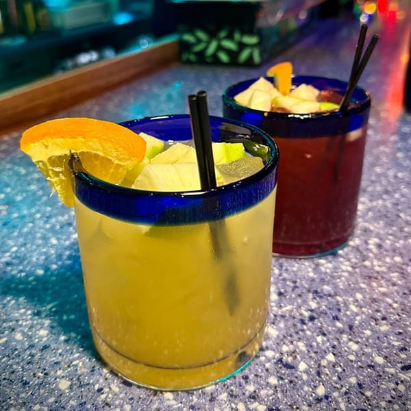 Two colorful Chilangos Restaurant drinks, one yellow and one red, garnished with fruit and straws.