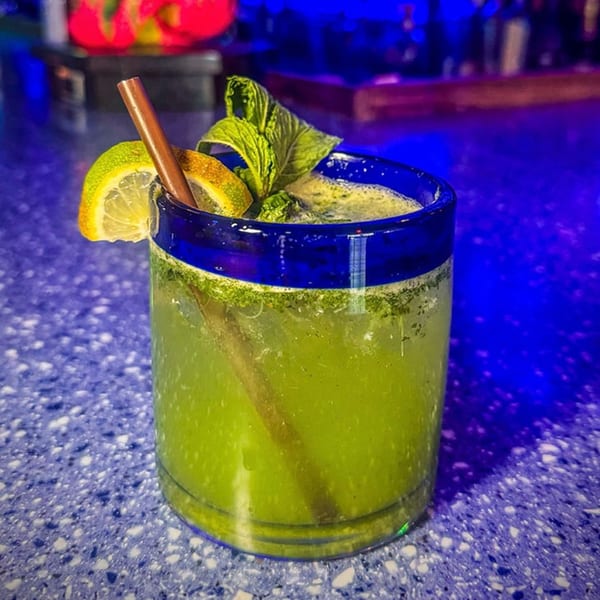 A vibrant green mojito with lime, mint, and a cinnamon stick in a blue-rimmed glass.